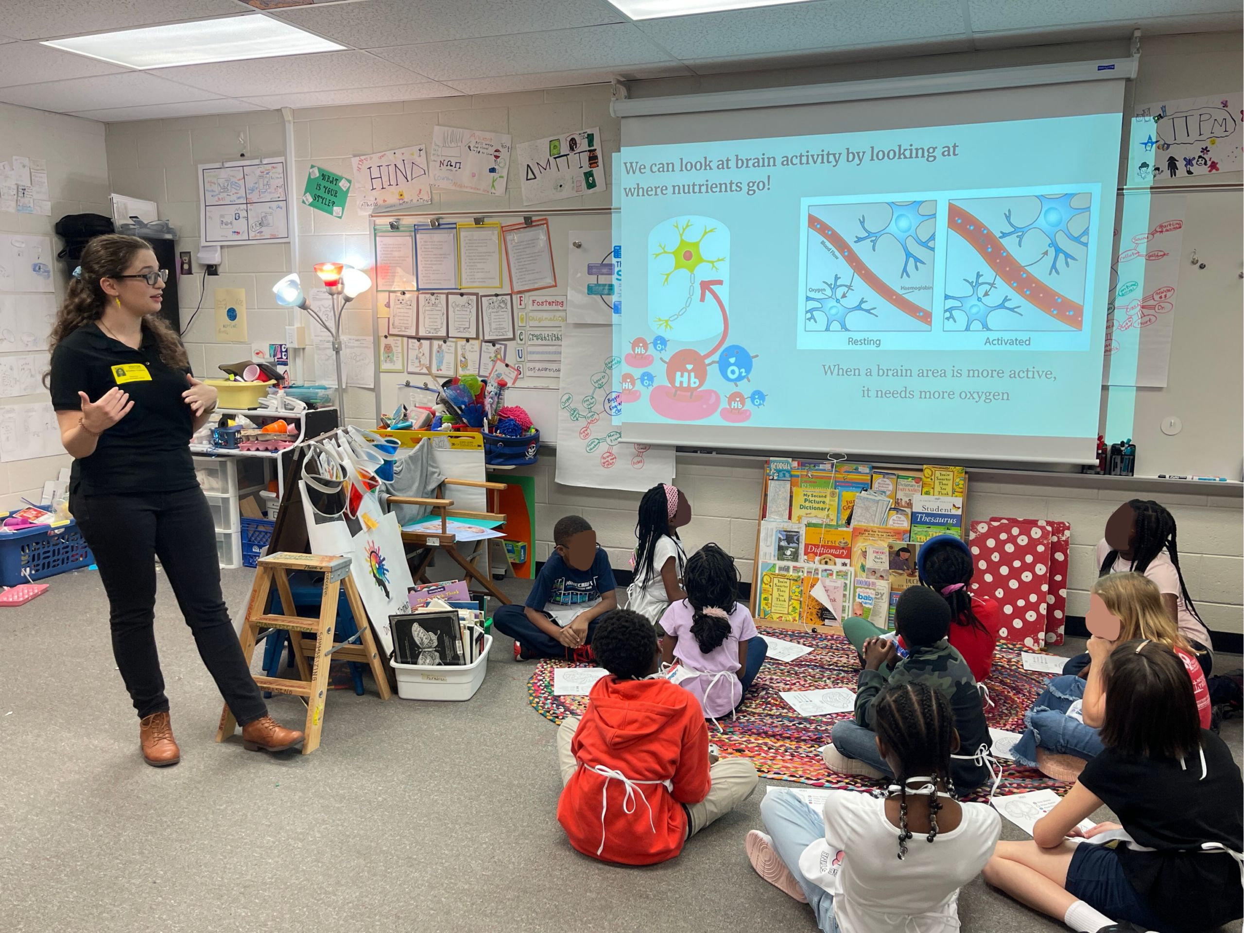 A woman explaining brain activity to a group of students sitting on a classroom floor. There is a projector displaying images related to brain function, with diagrams showing resting and activated brain areas. The classroom is filled with educational materials and decorations.